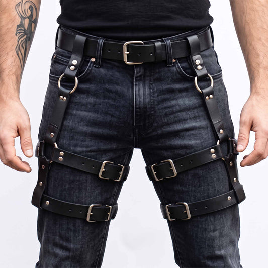 Front view of man wearing handmade black cowhide leather thigh harness with double roller buckle leg straps over black washed denim jeans