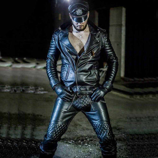 Full body front view of a man wearing a black cowhide leather biker jacket with padded sleeves, wide lapels, leather gloves, and leather pants.
