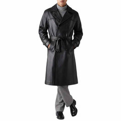 Full body view of a man wearing a black real cowhide leather double-breasted trench coat tied at the waist with grey trousers.