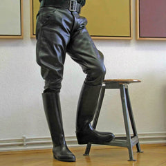 Black leather riding breeches with a black belt, worn with matching tall black leather riding boots, shown on a model standing on a metal stool in an indoor setting.