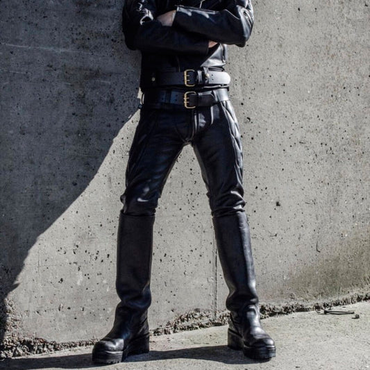 A person wearing black leather pants, a black leather jacket, and tall black leather boots, standing with arms crossed against a textured concrete wall.