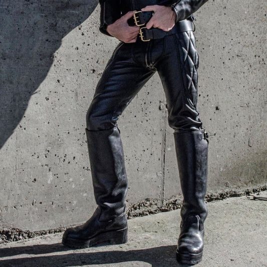 Person wearing black leather knee-high boots and pants against a concrete wall.