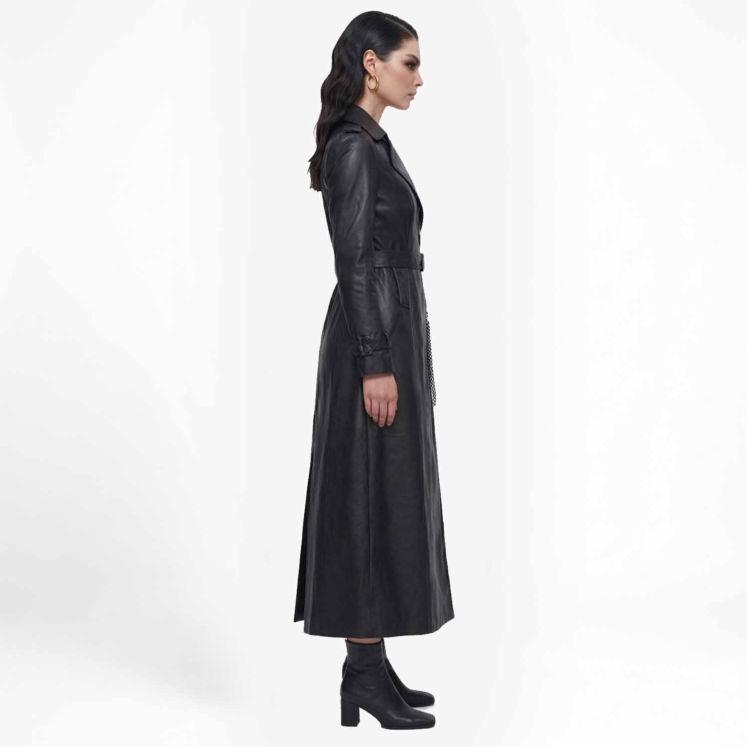 Side profile view of a woman wearing a long black leather trench coat highlighting its ankle-length silhouette.