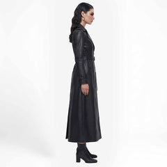 Side profile view of a woman wearing a long black leather trench coat highlighting its ankle-length silhouette.