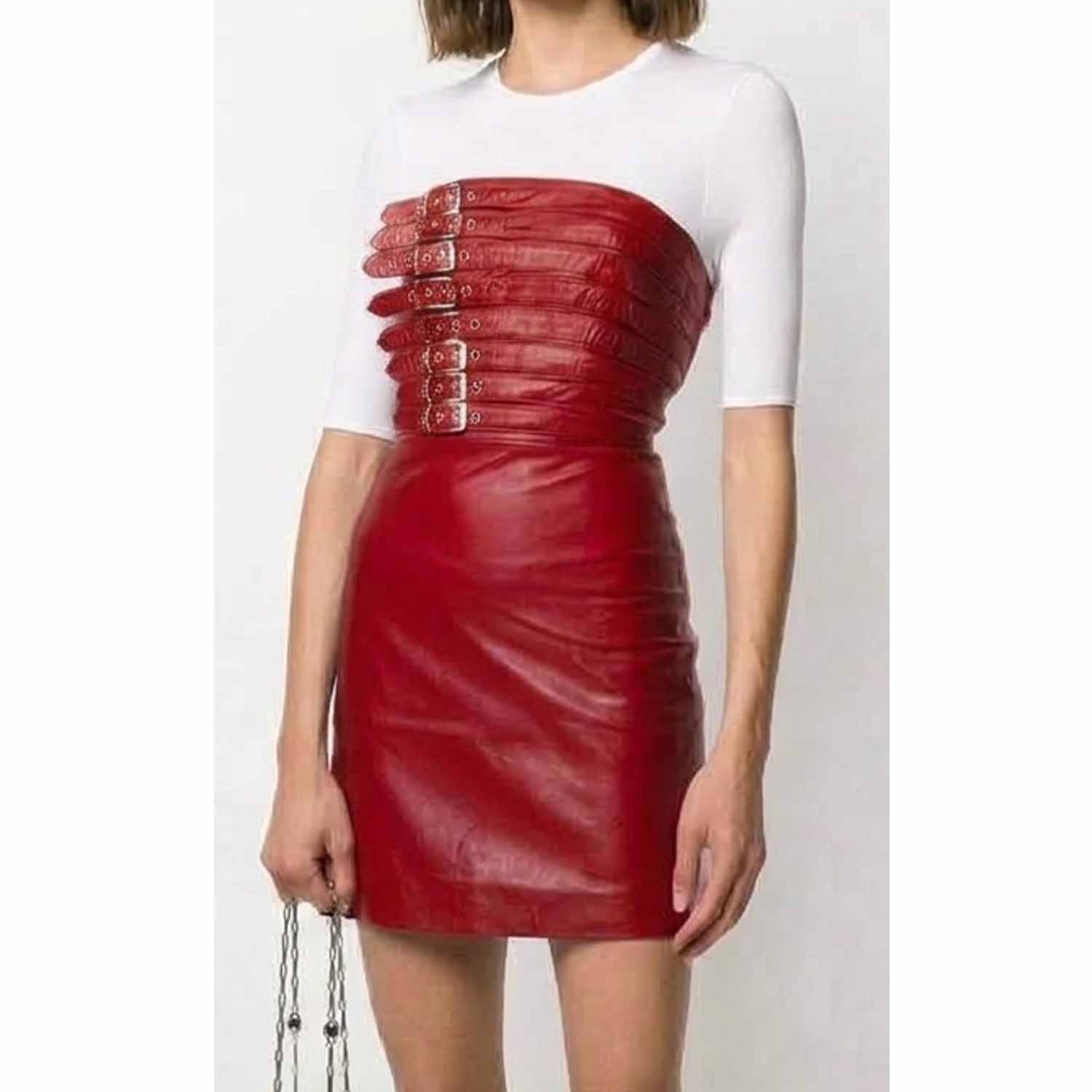A model wearing a red leather strapless mini skirt with a structured, corset-style bodice featuring multiple horizontal straps and silver-tone buckles, paired with a white short-sleeve top.